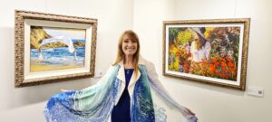 Jane Seymour standing between two of her paintings and modeling one of the scarves she design